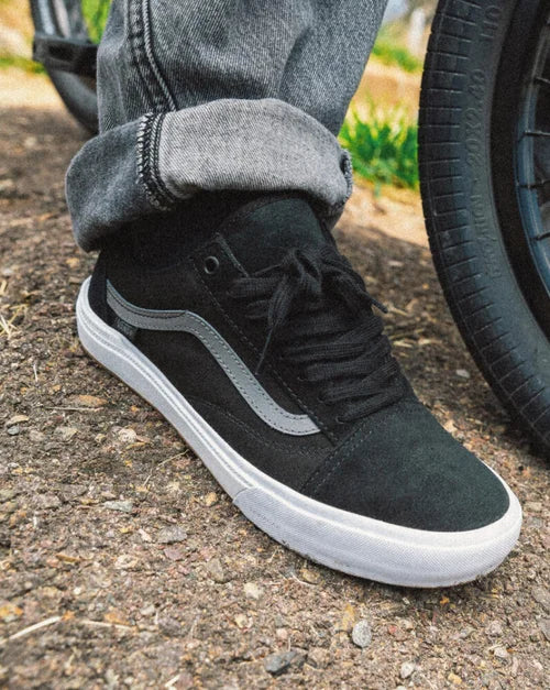 Black BMX Old Skool skate shoe with grey stripe worn by person standing on gravel next to bicycle wheel